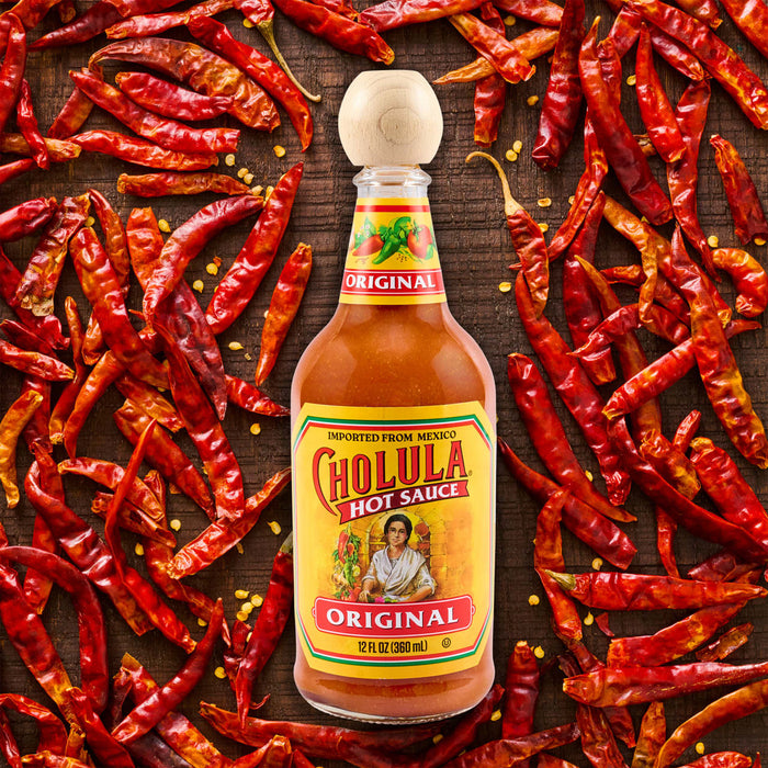 A bottle of Cholula Original Hot Sauce, featuring its iconic wooden cap, is centered on a dark wooden surface, surrounded by a vibrant array of dried red chili peppers and scattered seeds.