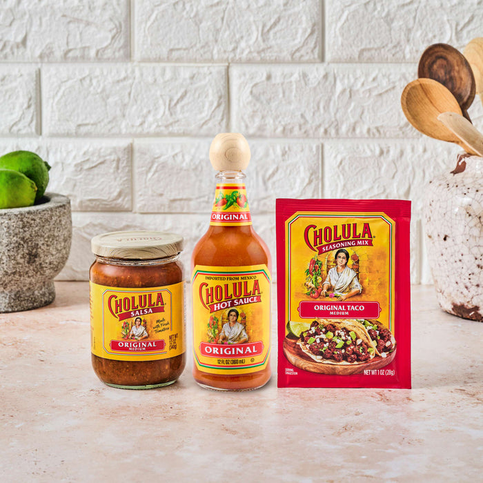 A jar of Cholula Salsa, a bottle of Cholula Original Hot Sauce, and a packet of Cholula Original Taco Seasoning Mix are displayed on a countertop next to a mortar and pestle with limes.