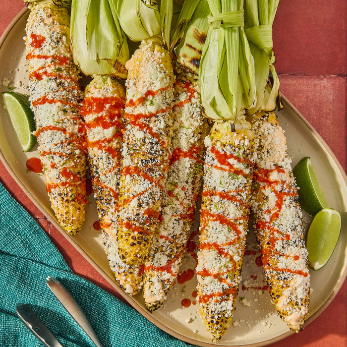 A platter of grilled corn on the cob, known as elotes, generously topped with crumbled cheese, herbs, and a vibrant drizzle of Cholula Original Hot Sauce, served with lime wedges.
