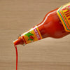 A bottle of Cholula Original Hot Sauce is tilted, with a stream of red sauce pouring out onto a light wooden surface.