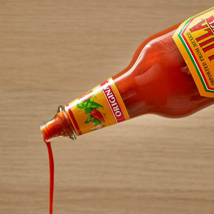 A bottle of Cholula Original Hot Sauce is tilted, with a stream of red sauce pouring out onto a light wooden surface.