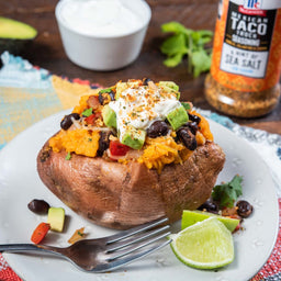 A baked sweet potato topped with black beans, avocado, sour cream, and spices, with a lime wedge and a fork on a white plate.