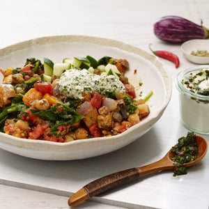 A vibrant bowl of vegetable and chickpea stew topped with herbed cheese and diced cucumber, with an eggplant, a red chili, and a small bowl containing Mccormick Gourmet Fennel Seed Organic in the background, alongside a jar of creamy dip and a wooden spoon with green pesto.