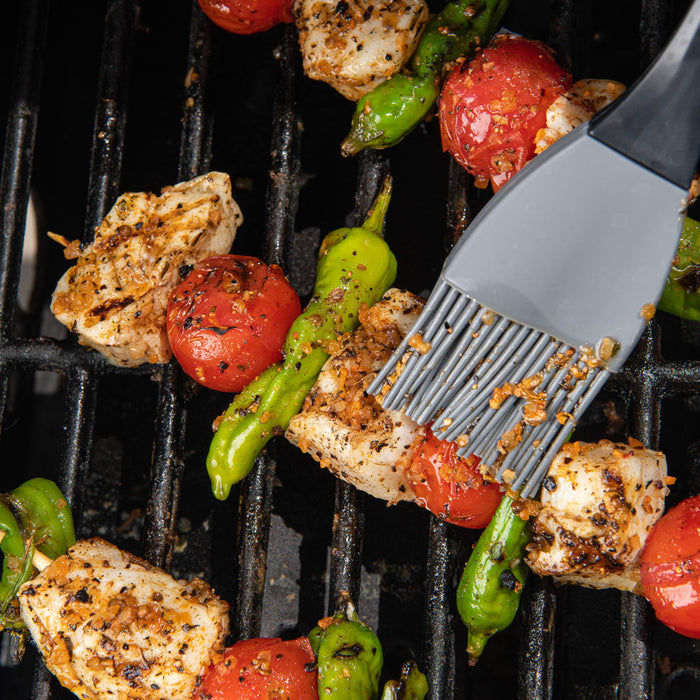 A basting brush applies Mccormick Grill Mates Roasted Garlic &amp;Herb Seasoning to chicken, tomatoes, and green peppers grilling on dark grates.
