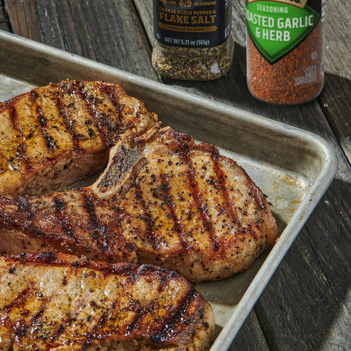 Three grilled pork chops seasoned with McCormick Grilling Coarse Black Pepper &amp; Flake Salt and Roasted Garlic &amp; Herb seasoning sit on a metal tray on a wooden surface.