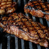 Three juicy steaks, seasoned with Mccormick Grill Mates Montreal Steak Seasoning, cooking on a hot grill with visible grill marks.