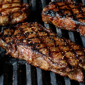 Three juicy steaks, seasoned with Mccormick Grill Mates Montreal Steak Seasoning, cooking on a hot grill with visible grill marks.
