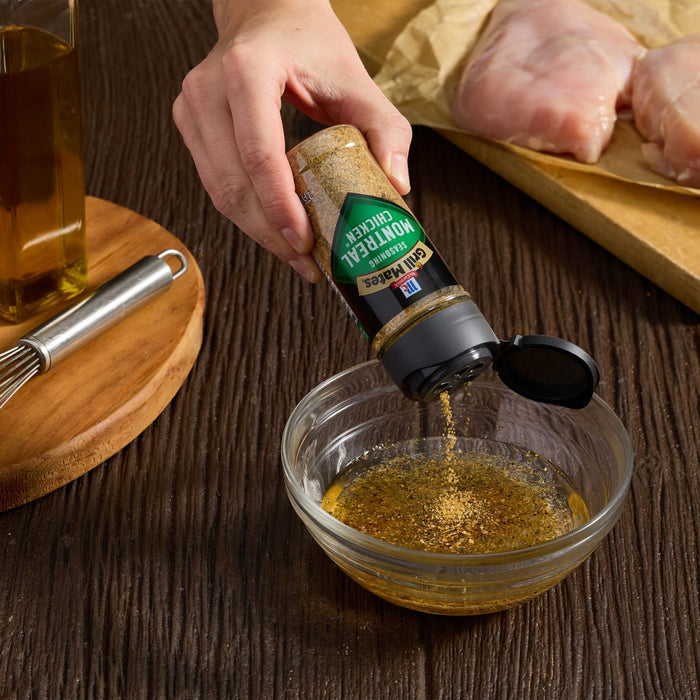 A person is adding McCormick's Montreal Chicken seasoning to a bowl of marinade next to raw chicken breasts.