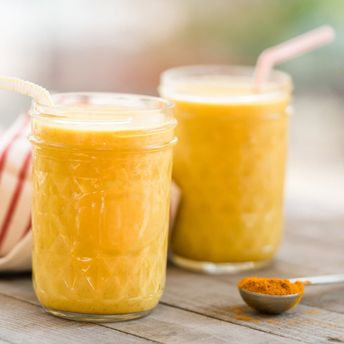Two glasses of yellow smoothie with a spoon of turmeric powder on a wooden table.