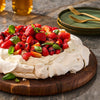 A pavlova dessert topped with fresh berries and basil sits on a wooden board, suggesting a light and refreshing summer treat.