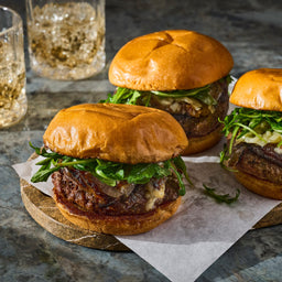 Three gourmet burgers with arugula and melted cheese are displayed on a wooden board, suggesting a high-end dining experience.