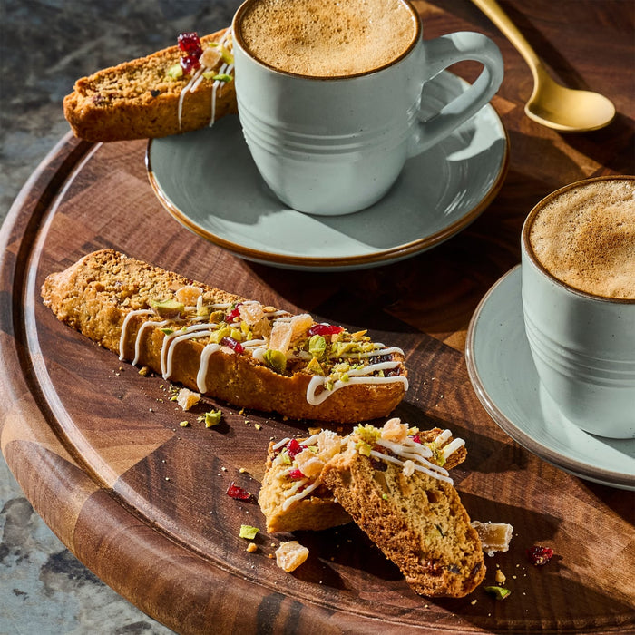 A wooden tray features biscotti drizzled with white icing and topped with pistachios and pomegranate seeds, alongside two cups of coffee.