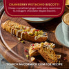 McCormick's cranberry pistachio biscotti, featuring crystallized ginger and chocolate drizzle, is displayed on a wooden board next to a cup of coffee.