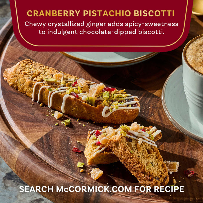 McCormick's cranberry pistachio biscotti, featuring crystallized ginger and chocolate drizzle, is displayed on a wooden board next to a cup of coffee.