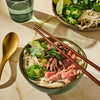 A bowl of Vietnamese Pho with beef, noodles, jalapenos, and herbs is presented with chopsticks and a golden spoon, suggesting a delicious and authentic culinary experience.