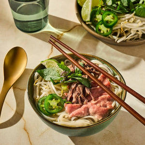 A bowl of Vietnamese Pho with beef, noodles, jalapenos, and herbs is presented with chopsticks and a golden spoon, suggesting a delicious and authentic culinary experience.