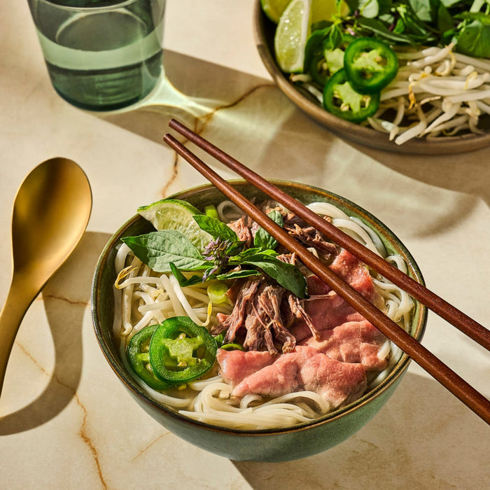 A bowl of Vietnamese Pho with beef, noodles, jalapenos, and herbs is presented with chopsticks and a golden spoon, suggesting a delicious and authentic culinary experience.