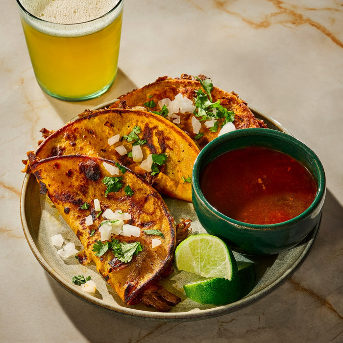 A plate of crispy birria tacos with a side of consommé, lime wedges, and a glass of beer.