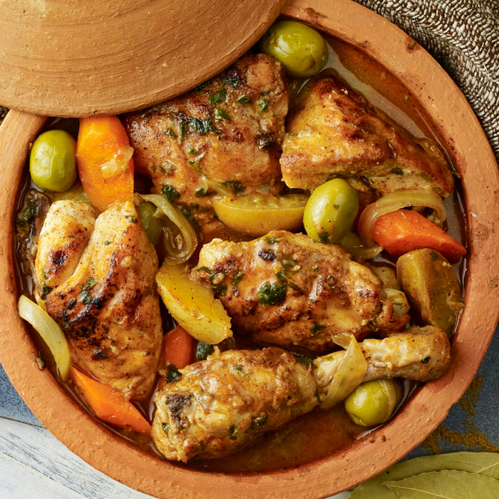 A close-up of a clay pot filled with chicken, carrots, potatoes, and olives.