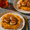 Two plates of seared scallops served over mashed sweet potatoes are presented on a textured surface, suggesting a gourmet dining experience.