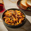 A bowl of pappardelle pasta with a rich meat sauce, accompanied by bread and a glass of red wine, suggesting a hearty and delicious meal.
