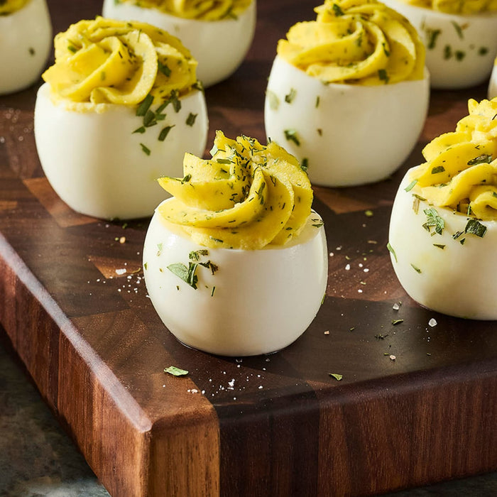A platter of deviled eggs, piped with a creamy yellow filling and garnished with herbs, sits on a wooden cutting board.