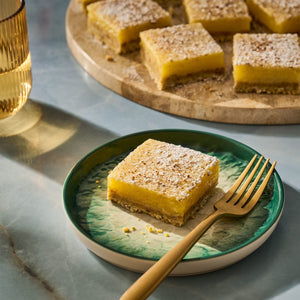 A lemon bar dusted with powdered sugar sits on a green plate with a gold fork, with more lemon bars on a wooden platter in the background.