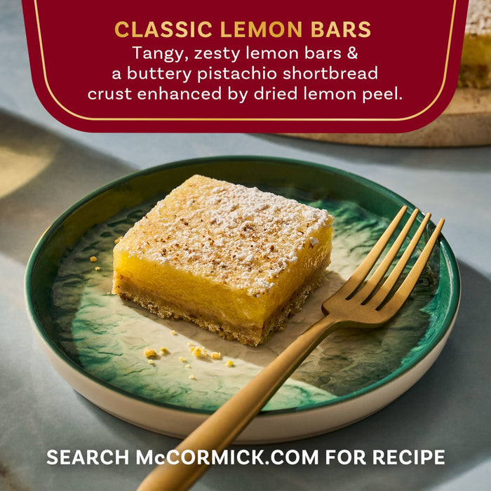 A close-up of a McCormick Classic Lemon Bar on a decorative plate with a fork, highlighting its tangy lemon filling and pistachio shortbread crust.
