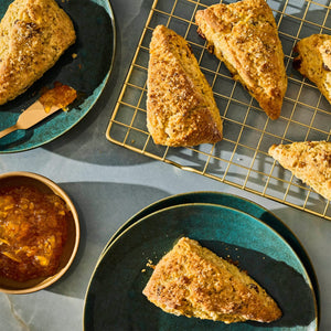 A top-down shot of golden brown scones arranged on teal plates and a gold wire rack, accompanied by a bowl of orange marmalade, showcasing a delicious breakfast or brunch spread.