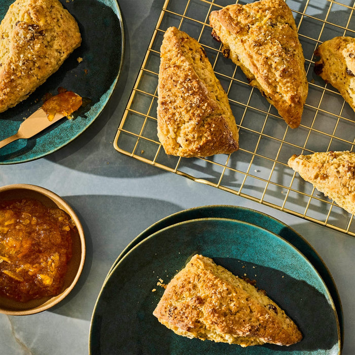 A top-down shot of golden brown scones arranged on teal plates and a gold wire rack, accompanied by a bowl of orange marmalade, showcasing a delicious breakfast or brunch spread.