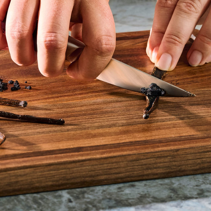 A person is slicing a vanilla bean on a wooden cutting board to extract the vanilla seeds.