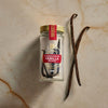A jar of Nielsen-Massey Madagascar Vanilla Beans sits next to two loose vanilla beans.
