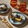 Two slices of lemon tart topped with whipped cream and berries are displayed on plates next to two glasses of amber liquid, suggesting a refreshing dessert pairing.