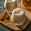 Two glasses of creamy eggnog topped with whipped cream and a sprinkle of spice, served with cinnamon sticks and a glass of whiskey.