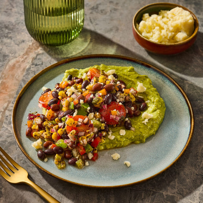 A vibrant plate features a bed of green puree topped with a colorful black bean and corn salsa, accompanied by a side of crumbled cheese.