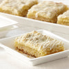 A close-up of a lemon bar with a white glaze on top, sitting on a white plate.
