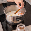 A person adds a cinnamon stick to a pot of milk and carrots on a stovetop, likely preparing a spiced beverage or dessert.