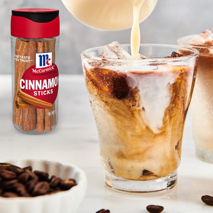 A glass of iced coffee with cream being poured in, alongside a container of McCormick Cinnamon Sticks and coffee beans.