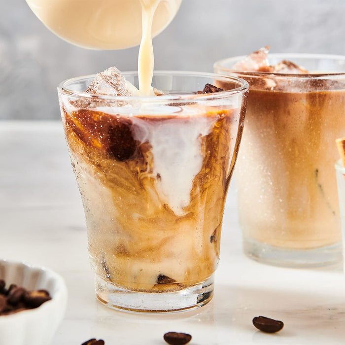 A refreshing iced coffee drink is being prepared with milk being poured into a glass filled with ice and coffee.