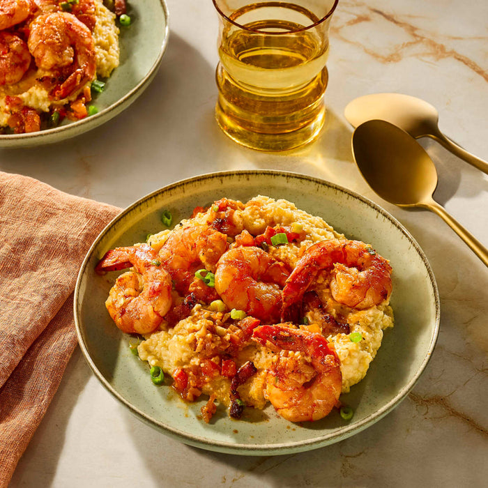 A plate of shrimp and grits is presented with a glass of white wine and gold utensils, suggesting a gourmet dining experience.