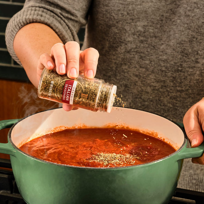 A person adds McCormick Italian Herb Seasoning to a pot of simmering tomato sauce.
