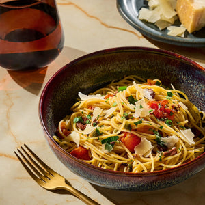 A bowl of spaghetti with tomatoes, herbs, and parmesan cheese is presented with a glass of wine and a side of parmesan shavings.