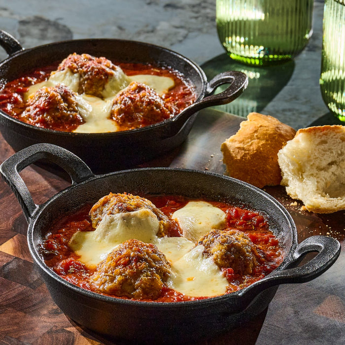 Two small cast iron skillets filled with meatballs in tomato sauce topped with melted cheese, served with biscuits.