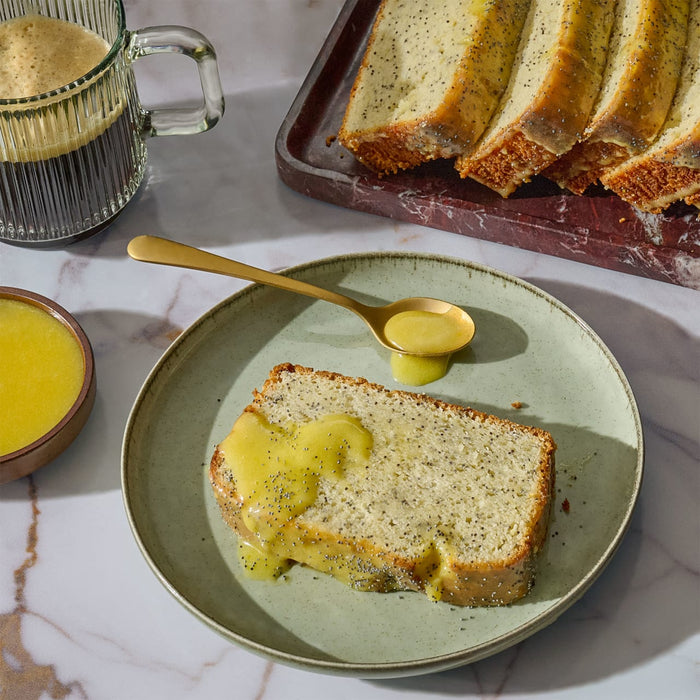 A slice of banana bread is drizzled with a bright yellow sauce, served on a plate with a spoon, alongside a mug of dark liquid and a tray of more bread slices.