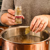 A person adds bay leaves from a spice jar labeled Bay Leaves to a large pot of simmering liquid.