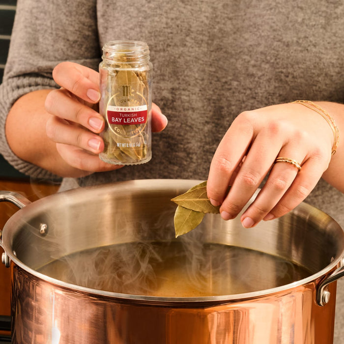 A person adds bay leaves from a spice jar labeled Bay Leaves to a large pot of simmering liquid.