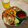A plate of crispy tacos with a side of consommé, lime wedges, and a glass of beer, showcasing a delicious and inviting meal.