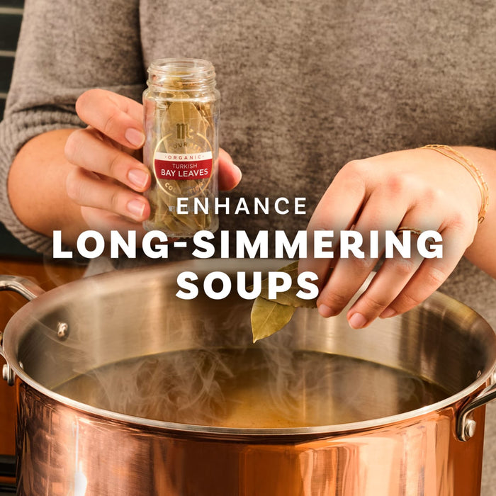 A person adds bay leaves from a McCormick spice jar to a large pot of simmering soup to enhance the flavor.