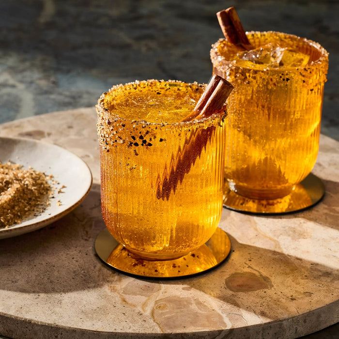 Two festive cocktails garnished with cinnamon sticks and a sugared rim sit on a wooden board, suggesting a seasonal or holiday theme.