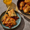 A plate of braised chicken with beans and bread is presented alongside a cast iron skillet containing more of the same dish, suggesting a hearty and flavorful meal.